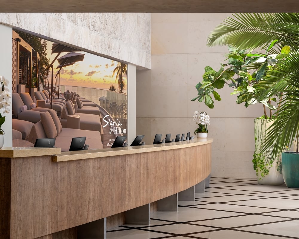 A curved wooden reception desk with small monitors stands before a large screen displaying a sunset beach scene with lounge chairs in a modern, plant-filled lobby. Text: Signia by Hilton DIPLOMAT BEACH.
