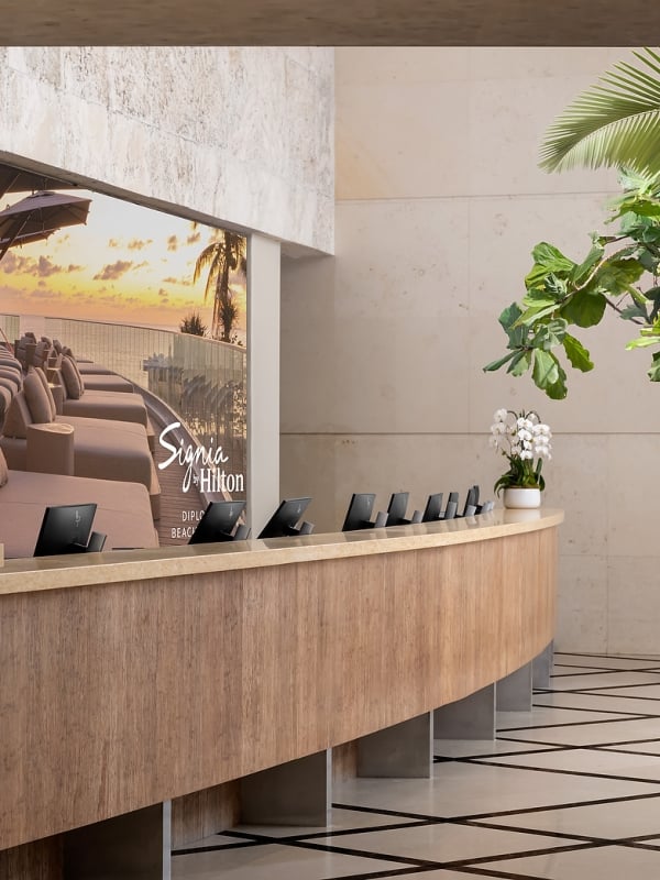 A curved wooden reception desk with small monitors stands before a large screen displaying a sunset beach scene with lounge chairs in a modern, plant-filled lobby. Text: Signia by Hilton DIPLOMAT BEACH.