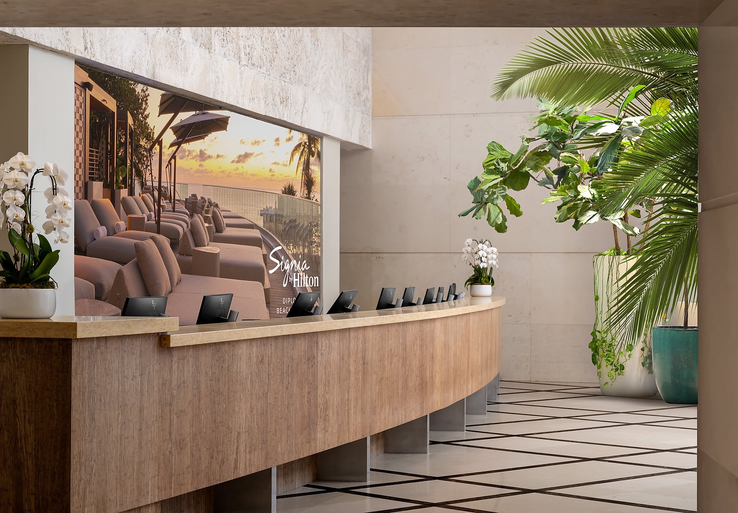A curved wooden reception desk with small monitors stands before a large screen displaying a sunset beach scene with lounge chairs in a modern, plant-filled lobby. Text: Signia by Hilton DIPLOMAT BEACH.