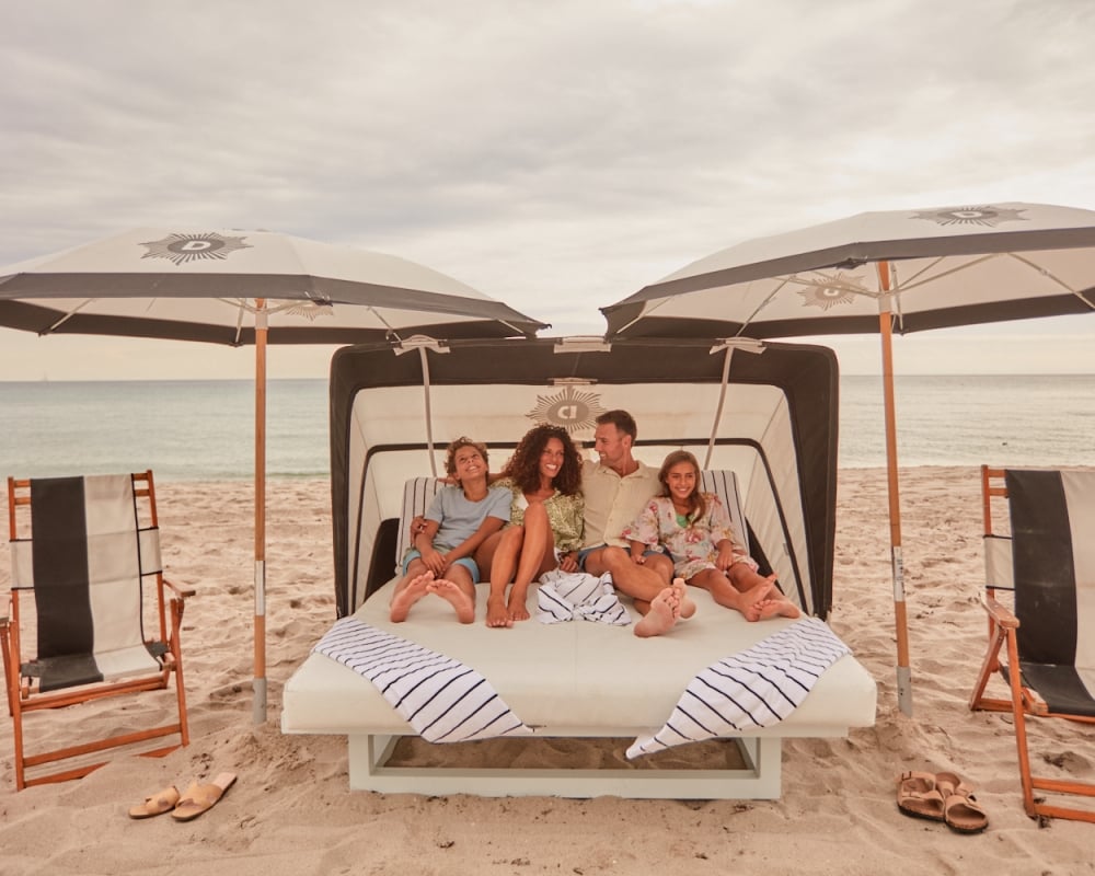 A family of four relaxes on a daybed under a black and white cabana (CJ) and umbrellas on a sandy beach with the ocean nearby.