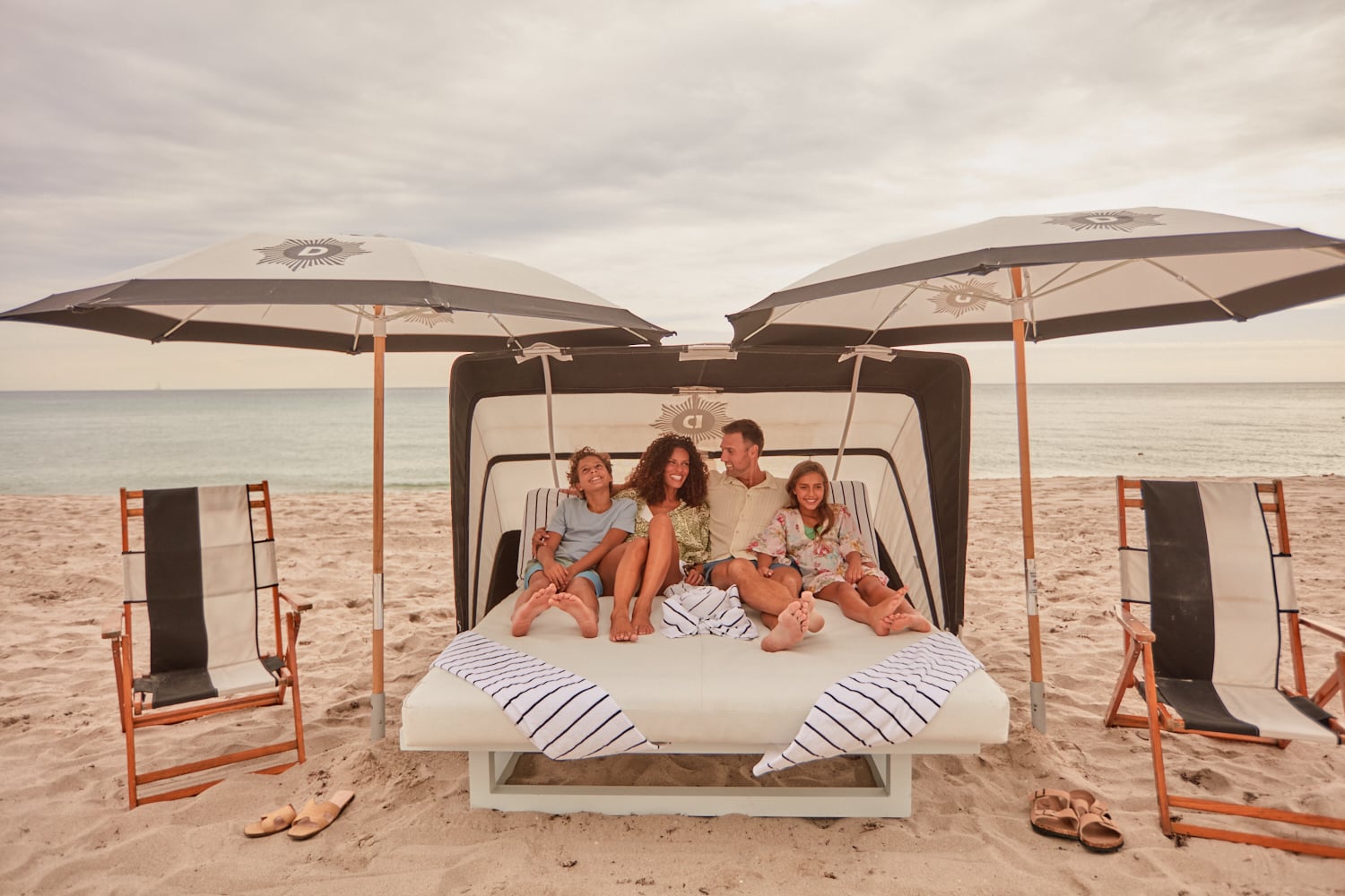 A family of four relaxes on a daybed under a black and white cabana (CJ) and umbrellas on a sandy beach with the ocean nearby.