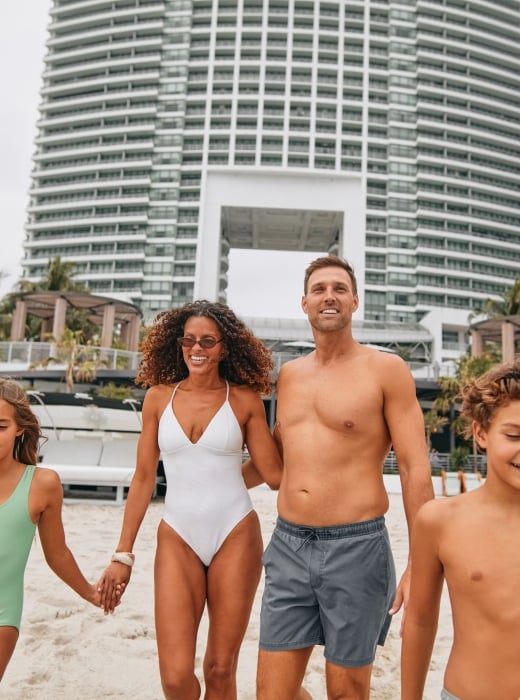 A smiling family of four walks hand-in-hand on a sandy beach, with tall white buildings and palm trees in the background.