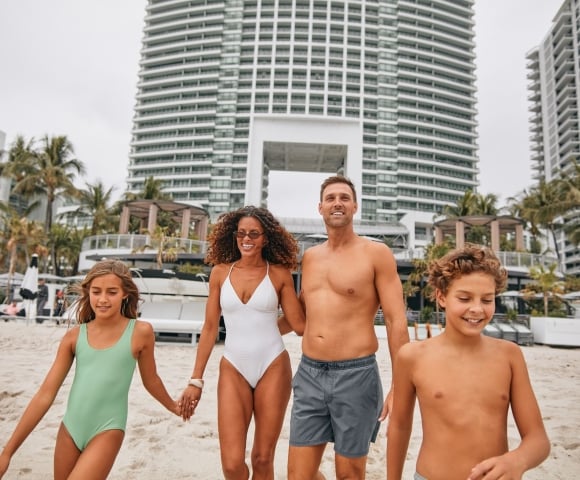 A smiling family of four walks hand-in-hand on a sandy beach, with tall white buildings and palm trees in the background.