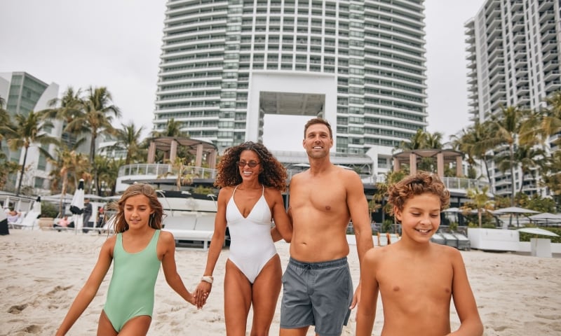 A smiling family of four walks hand-in-hand on a sandy beach, with tall white buildings and palm trees in the background.