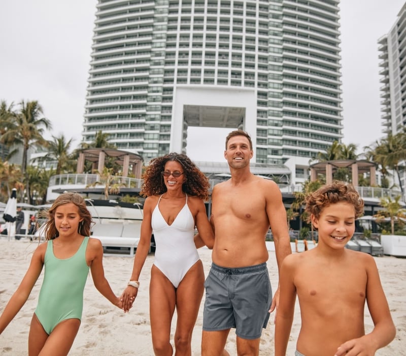 A smiling family of four walks hand-in-hand on a sandy beach, with tall white buildings and palm trees in the background.