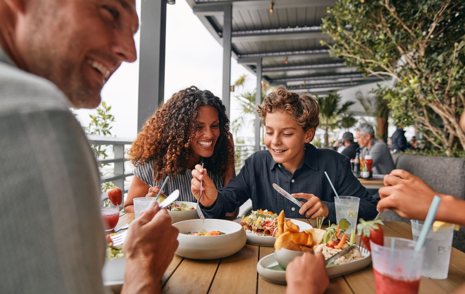 A smiling family, including a man, woman, and boy, enjoys a meal at an outdoor restaurant. They are eating various dishes and drinking from cups that say 'PLA BEACH BAR'.