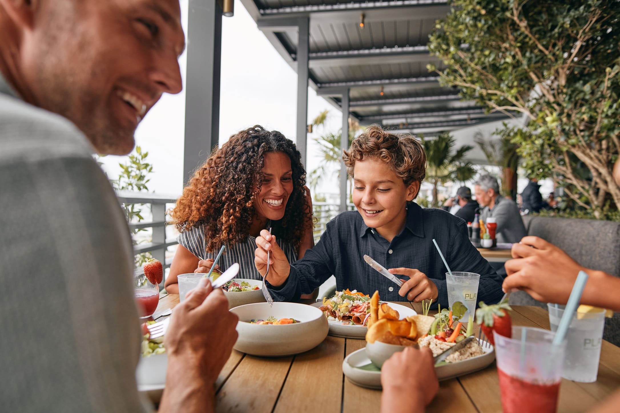 A smiling family, including a man, woman, and boy, enjoys a meal at an outdoor restaurant. They are eating various dishes and drinking from cups that say 'PLA BEACH BAR'.