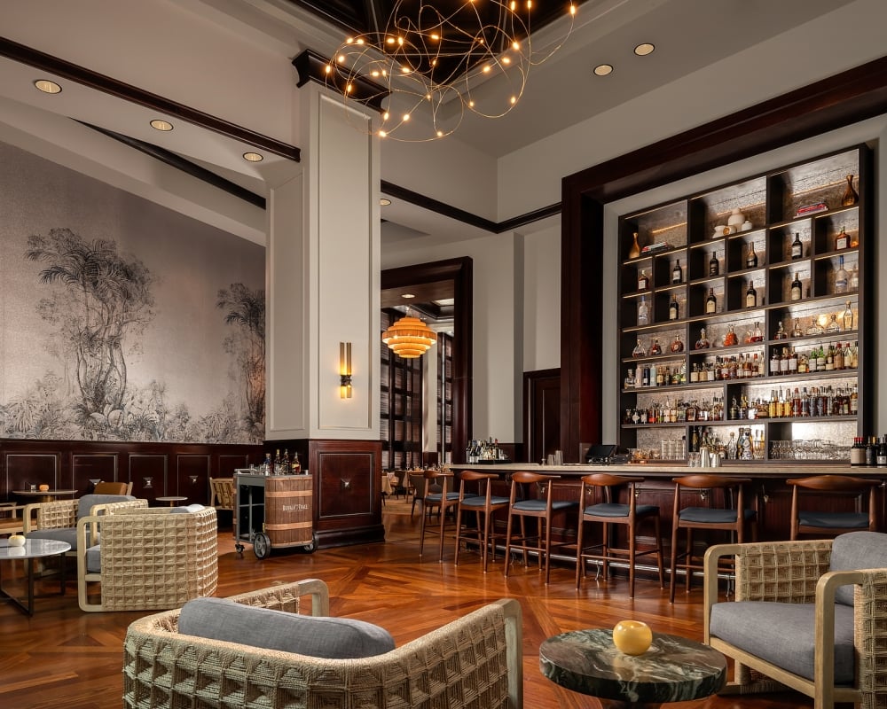 Elegant bar and lounge area with a long, well-stocked counter, various modern seating, patterned wooden floor, and botanical mural. A barrel cart reads: Buffalo Trace.