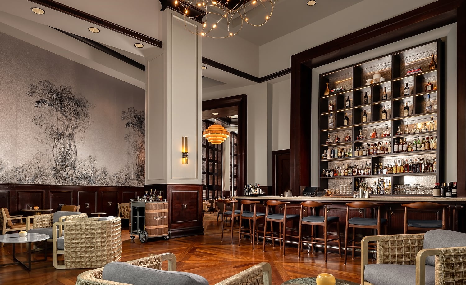 Elegant bar and lounge area with a long, well-stocked counter, various modern seating, patterned wooden floor, and botanical mural. A barrel cart reads: Buffalo Trace.