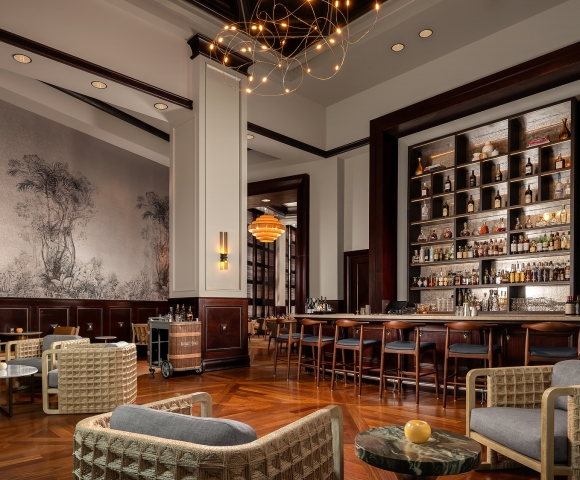 Elegant bar and lounge area with a long, well-stocked counter, various modern seating, patterned wooden floor, and botanical mural. A barrel cart reads: Buffalo Trace.