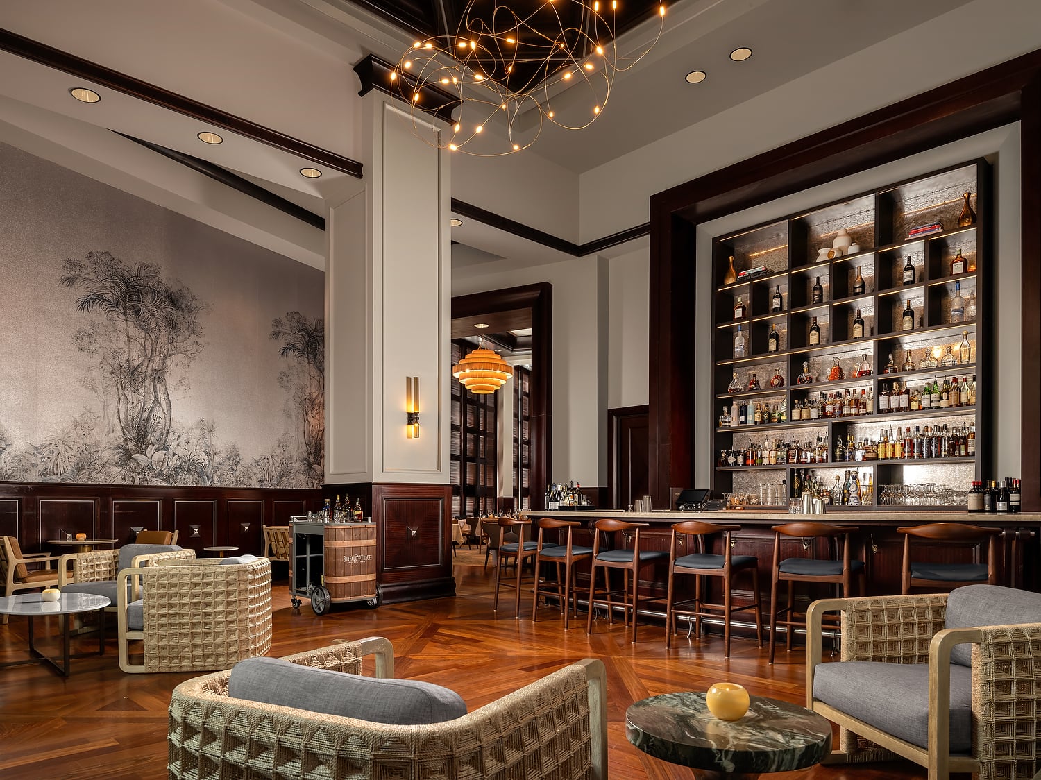 Elegant bar and lounge area with a long, well-stocked counter, various modern seating, patterned wooden floor, and botanical mural. A barrel cart reads: Buffalo Trace.