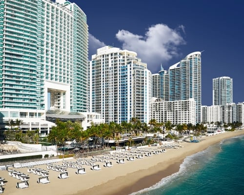 Modern high-rise buildings stand along a wide sandy beach, packed with white loungers, beside a turquoise ocean under a clear blue sky.