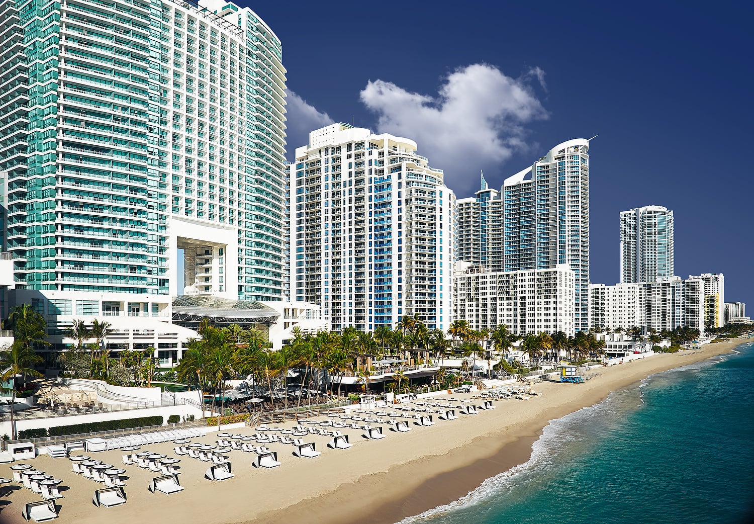 Modern high-rise buildings stand along a wide sandy beach, packed with white loungers, beside a turquoise ocean under a clear blue sky.