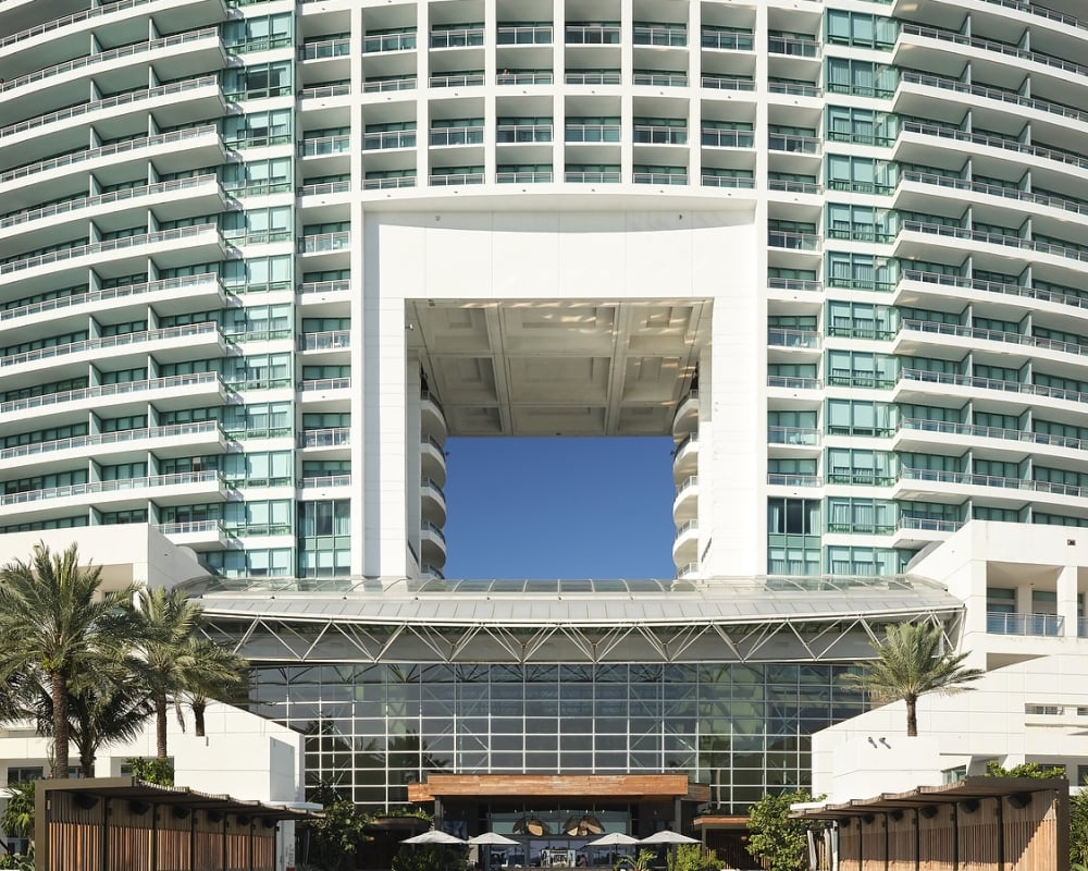 Curved high-rise building with a large central void overlooks a resort pool, surrounded by lounge chairs, cabanas, and palm trees under a blue sky. Text: 4 FT 3 IN