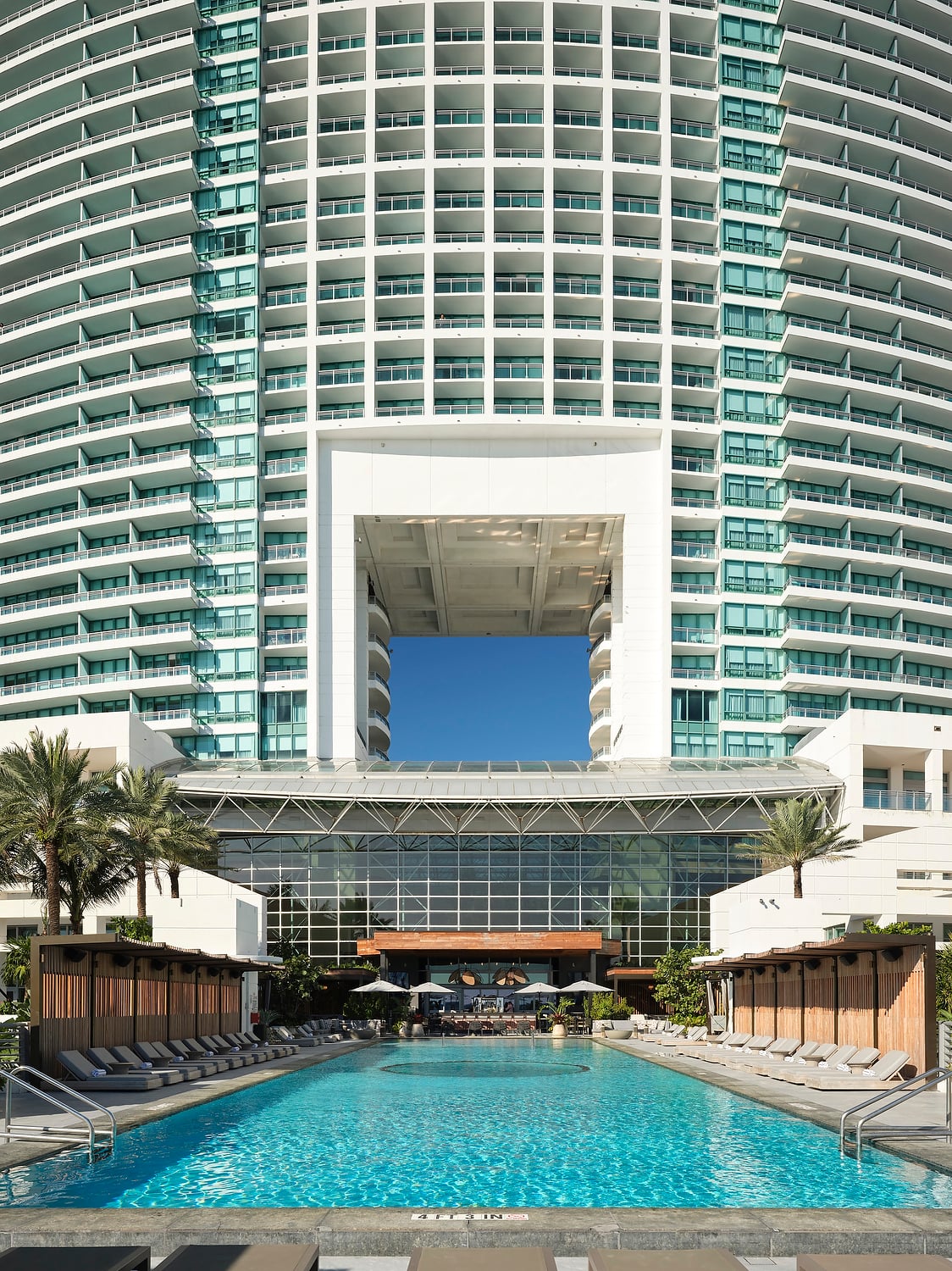 Curved high-rise building with a large central void overlooks a resort pool, surrounded by lounge chairs, cabanas, and palm trees under a blue sky. Text: 4 FT 3 IN