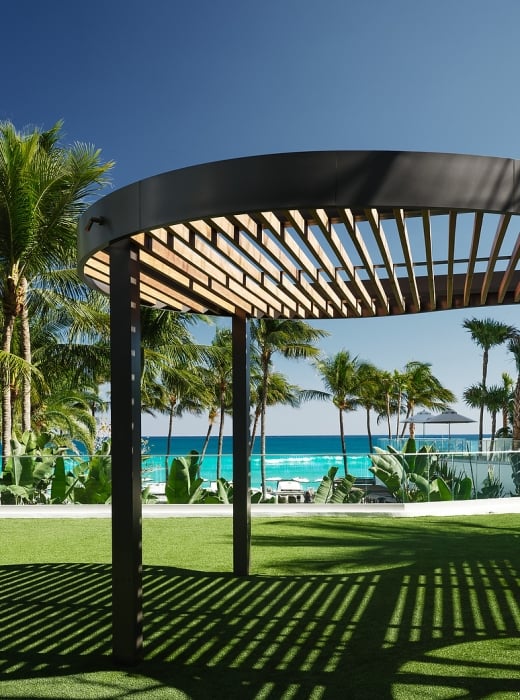 A curved, slatted pergola casts striped shadows on green artificial grass, with palm trees and a turquoise ocean under a clear blue sky in the background.