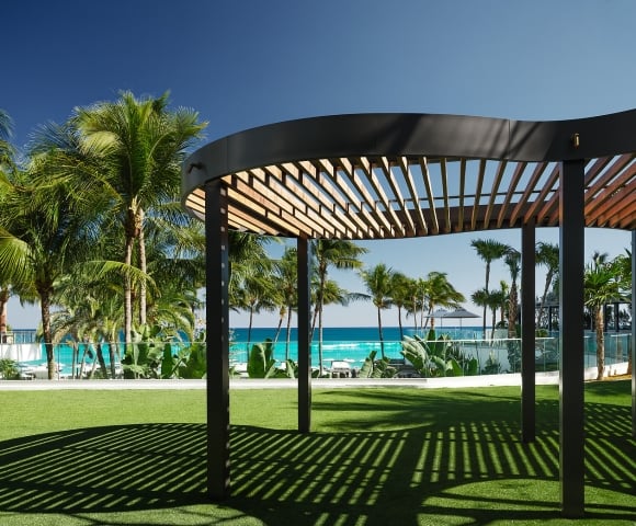 A curved, slatted pergola casts striped shadows on green artificial grass, with palm trees and a turquoise ocean under a clear blue sky in the background.