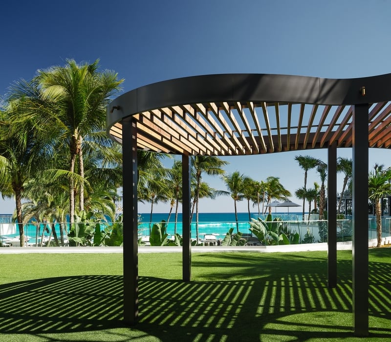 A curved, slatted pergola casts striped shadows on green artificial grass, with palm trees and a turquoise ocean under a clear blue sky in the background.