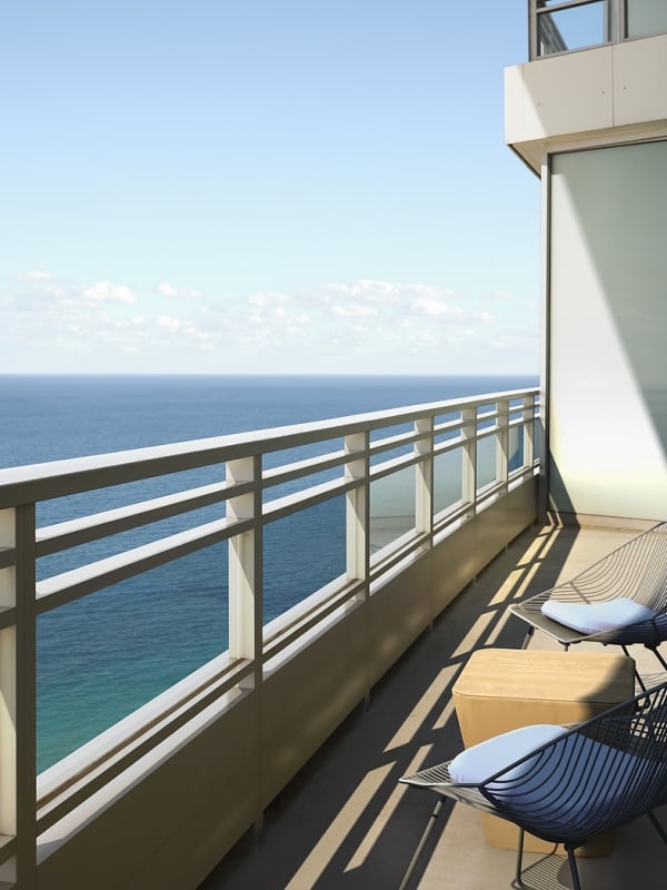 A high-rise balcony features two dark wireframe chairs with light blue cushions and a wooden table, overlooking a vast blue ocean under a clear sky.