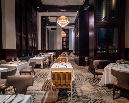 An elegant, long dining room with tables set for meals, featuring white tablecloths and tableware. Ornate chandeliers illuminate dark wood-paneled walls and a patterned carpet, creating a sophisticated ambiance.