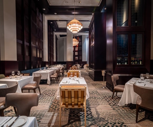 An elegant, long dining room with tables set for meals, featuring white tablecloths and tableware. Ornate chandeliers illuminate dark wood-paneled walls and a patterned carpet, creating a sophisticated ambiance.