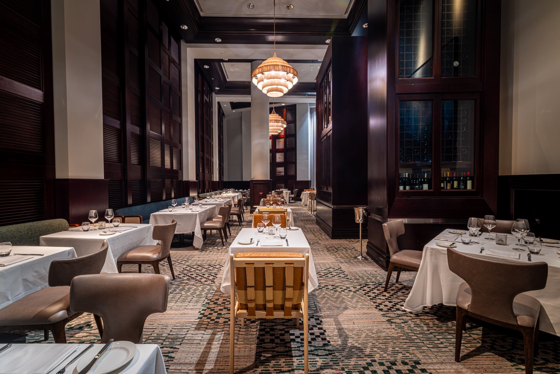 An elegant, long dining room with tables set for meals, featuring white tablecloths and tableware. Ornate chandeliers illuminate dark wood-paneled walls and a patterned carpet, creating a sophisticated ambiance.