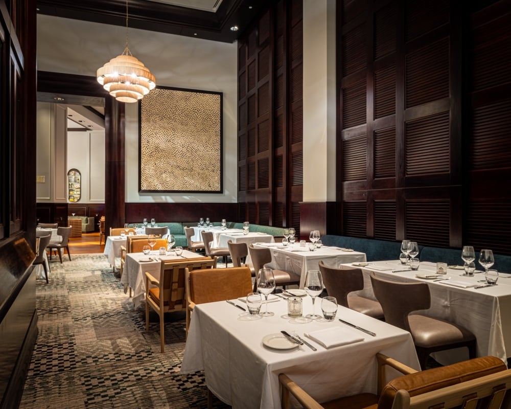 Restaurant dining room with white-clothed tables set with glassware and cutlery. Dark wood walls, unique chandelier, textured artwork, and patterned carpet. Text: EXIT.