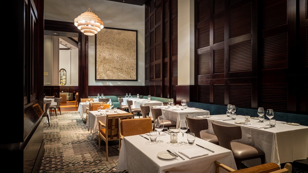 Restaurant dining room with white-clothed tables set with glassware and cutlery. Dark wood walls, unique chandelier, textured artwork, and patterned carpet. Text: EXIT.