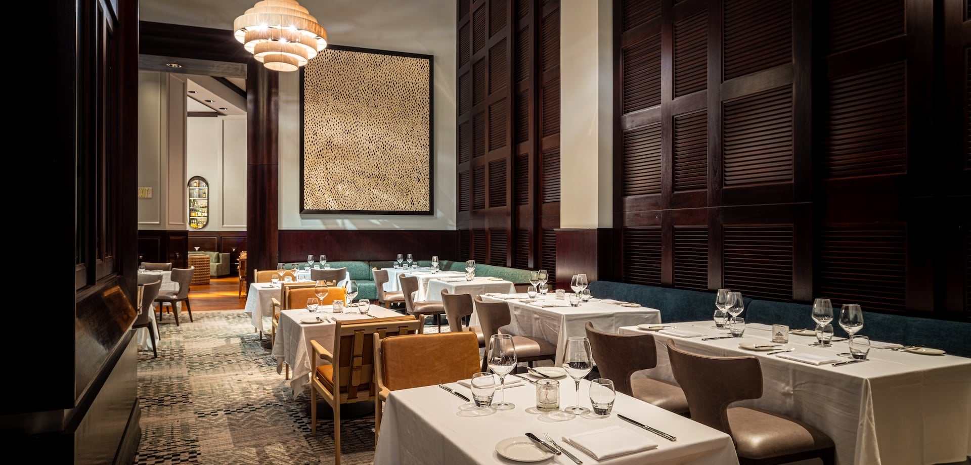 Restaurant dining room with white-clothed tables set with glassware and cutlery. Dark wood walls, unique chandelier, textured artwork, and patterned carpet. Text: EXIT.