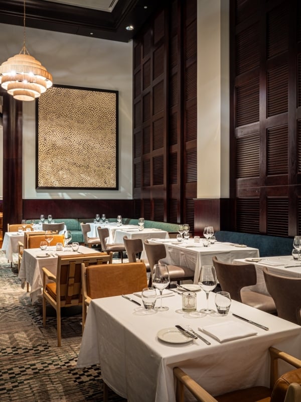 Restaurant dining room with white-clothed tables set with glassware and cutlery. Dark wood walls, unique chandelier, textured artwork, and patterned carpet. Text: EXIT.