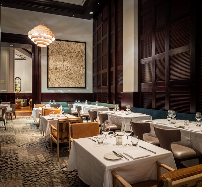 Restaurant dining room with white-clothed tables set with glassware and cutlery. Dark wood walls, unique chandelier, textured artwork, and patterned carpet. Text: EXIT.