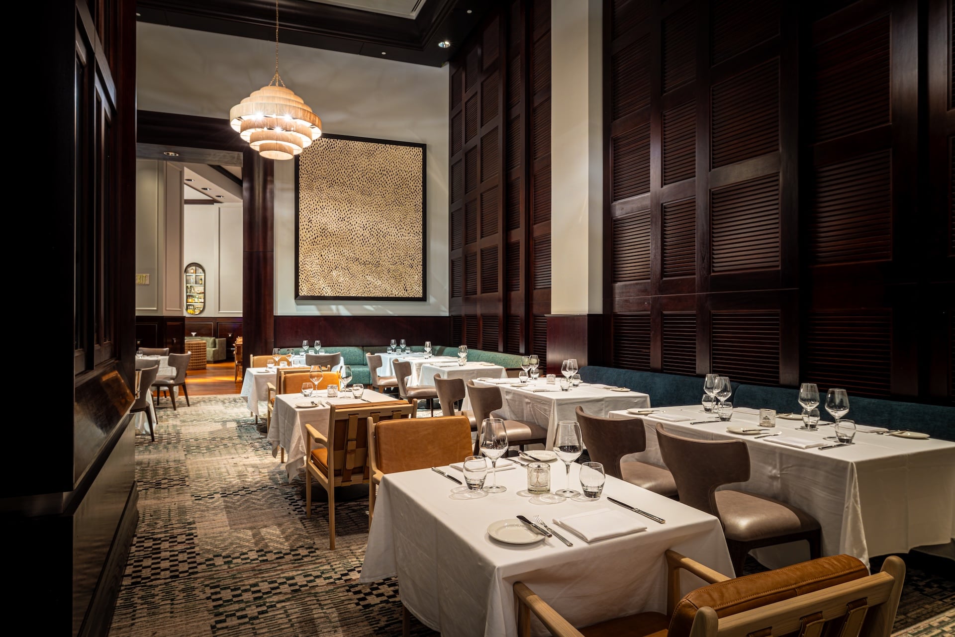 Restaurant dining room with white-clothed tables set with glassware and cutlery. Dark wood walls, unique chandelier, textured artwork, and patterned carpet. Text: EXIT.