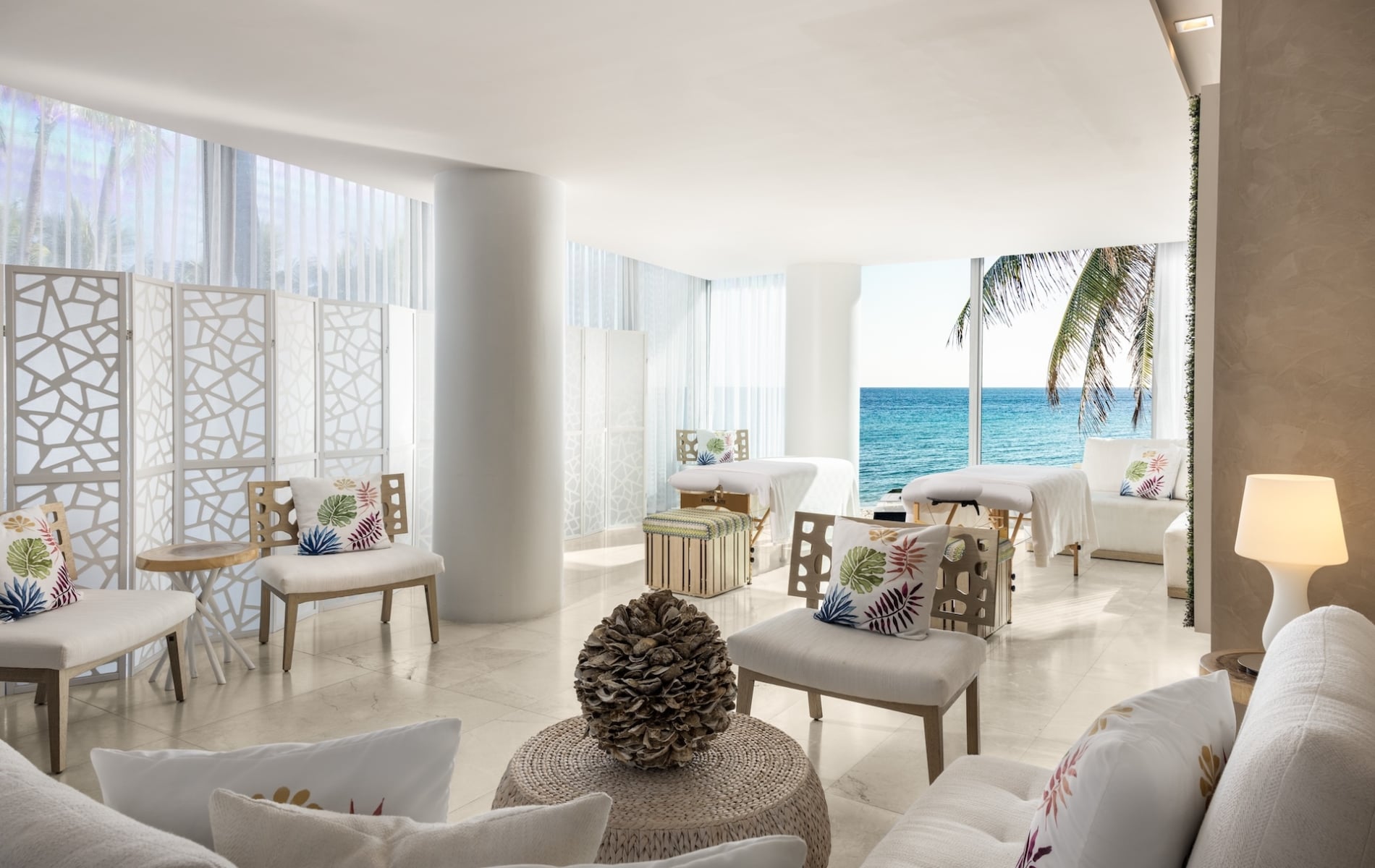 A brightly lit spa room features multiple massage tables and seating areas. Large windows showcase a vibrant blue ocean and a palm tree.