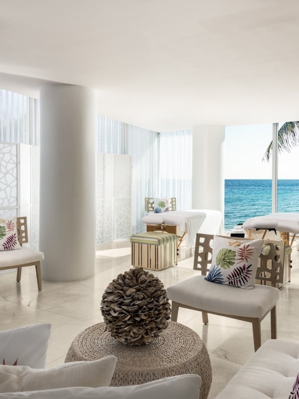 A brightly lit spa room features multiple massage tables and seating areas. Large windows showcase a vibrant blue ocean and a palm tree.