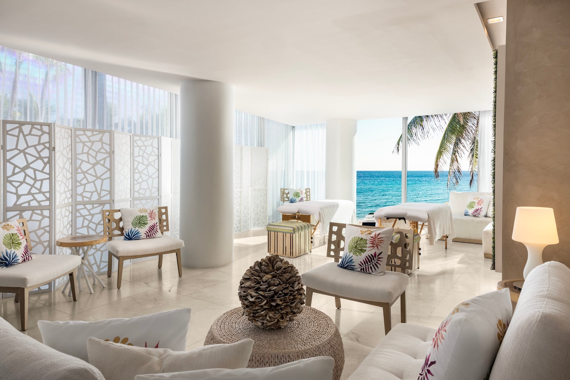 A brightly lit spa room features multiple massage tables and seating areas. Large windows showcase a vibrant blue ocean and a palm tree.