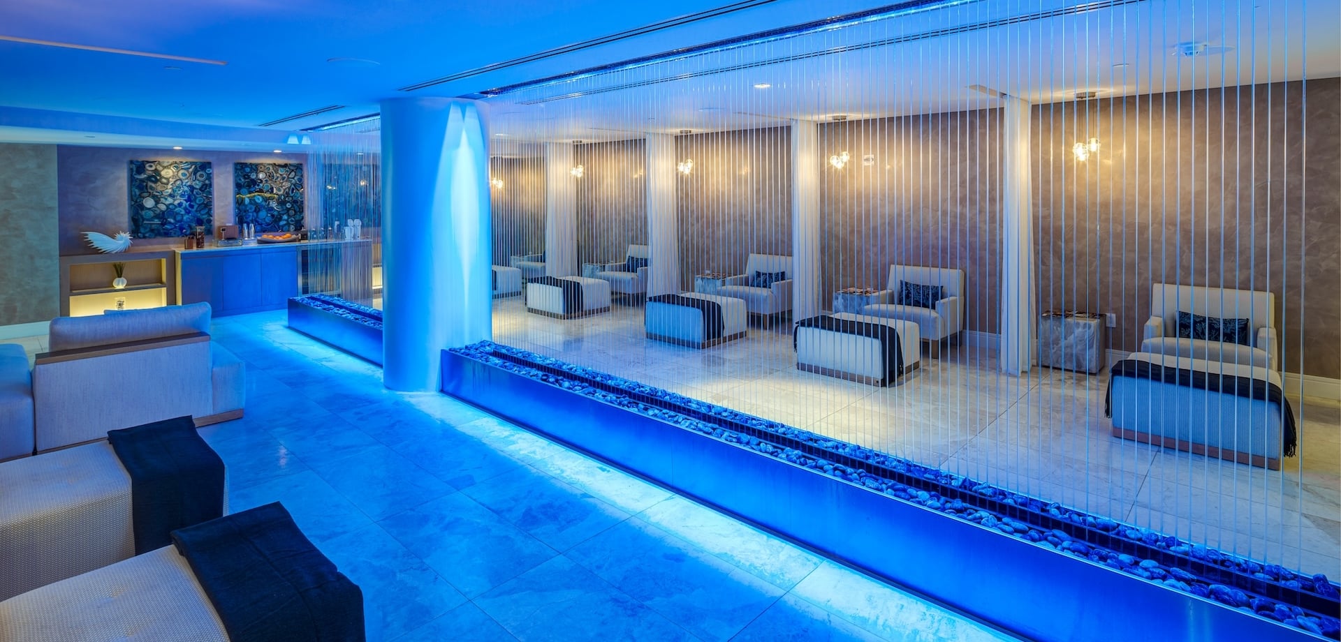 A modern spa lounge is illuminated by vibrant blue lighting, featuring a central pillar with a glowing waterfall effect and parallel rock-filled troughs. Seating areas and a beverage bar complete the serene environment.