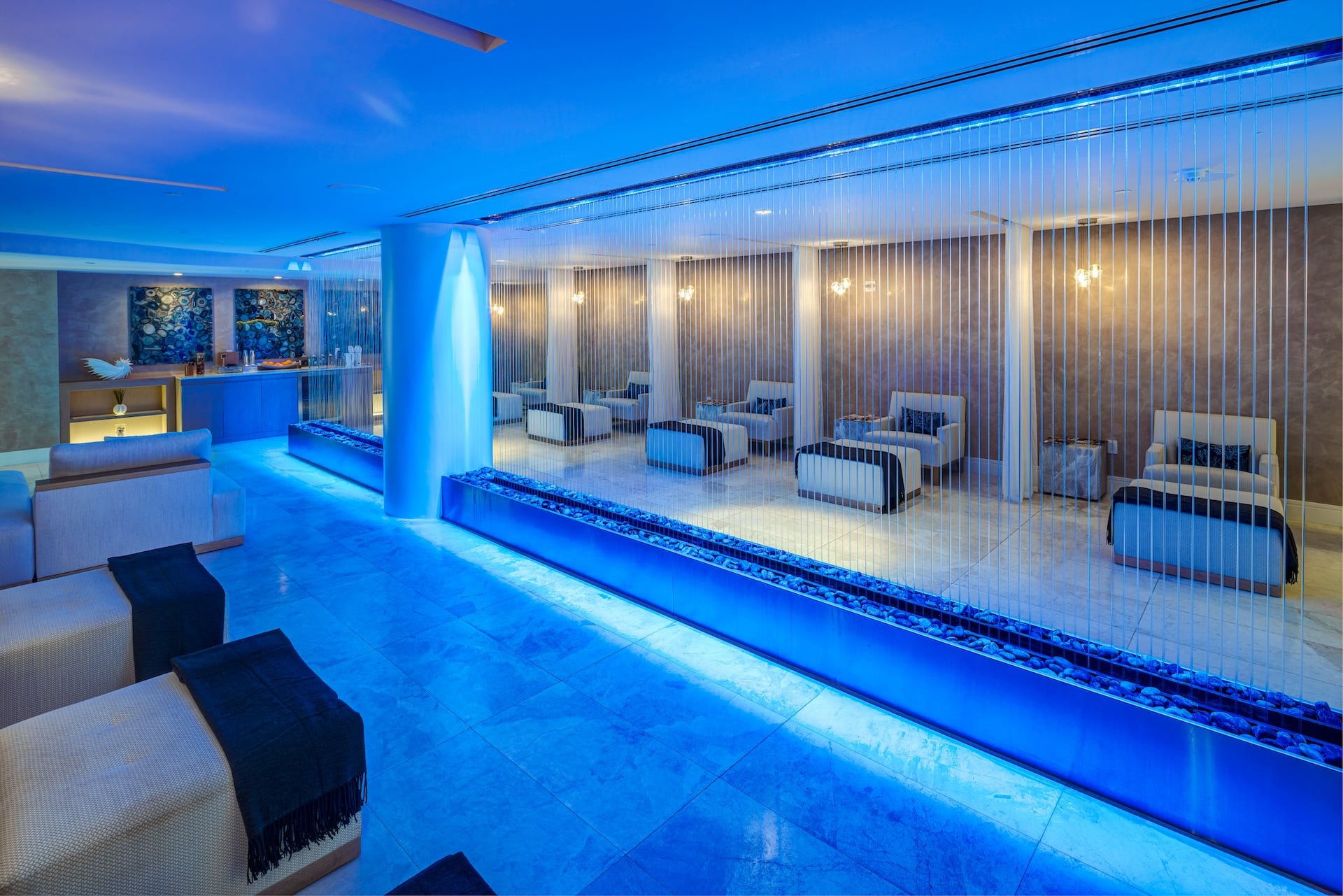 A modern spa lounge is illuminated by vibrant blue lighting, featuring a central pillar with a glowing waterfall effect and parallel rock-filled troughs. Seating areas and a beverage bar complete the serene environment.