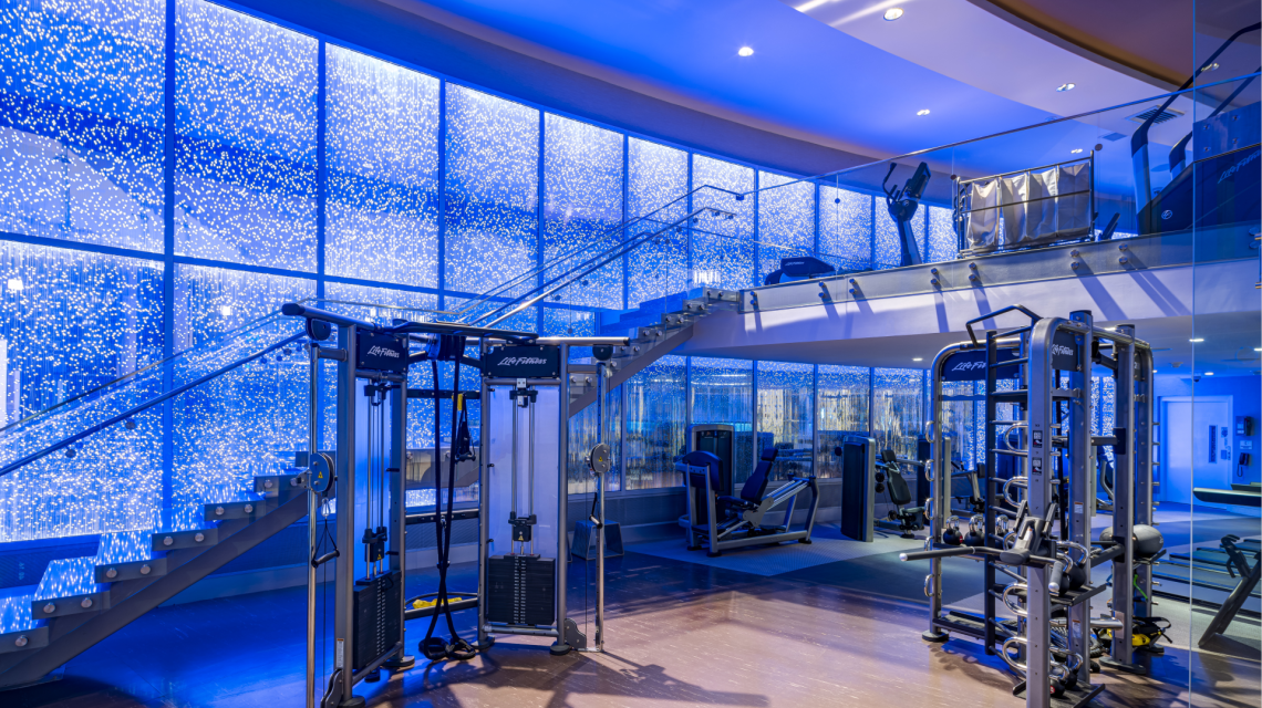 Life Fitness gym equipment fills a modern, multi-level fitness center featuring blue lighting, a glass staircase, and sparkling light-dotted walls. Text: Life Fitness