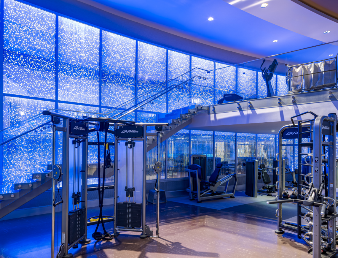 Life Fitness gym equipment fills a modern, multi-level fitness center featuring blue lighting, a glass staircase, and sparkling light-dotted walls. Text: Life Fitness