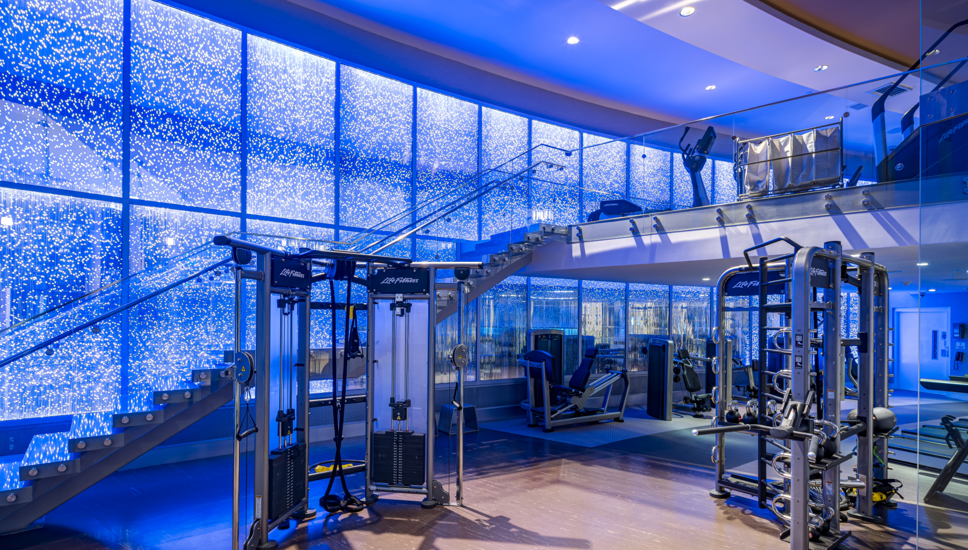Life Fitness gym equipment fills a modern, multi-level fitness center featuring blue lighting, a glass staircase, and sparkling light-dotted walls. Text: Life Fitness