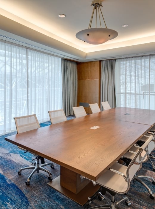 A large wooden conference table with modern chairs stands empty in a meeting room, surrounded by sheer white curtains and an abstract blue/orange carpet.