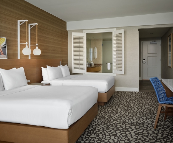 Two beds with white linens are in a hotel room. It features a wood wall, patterned carpet, a bathroom visible through open shutters, and a blue chair at a desk.