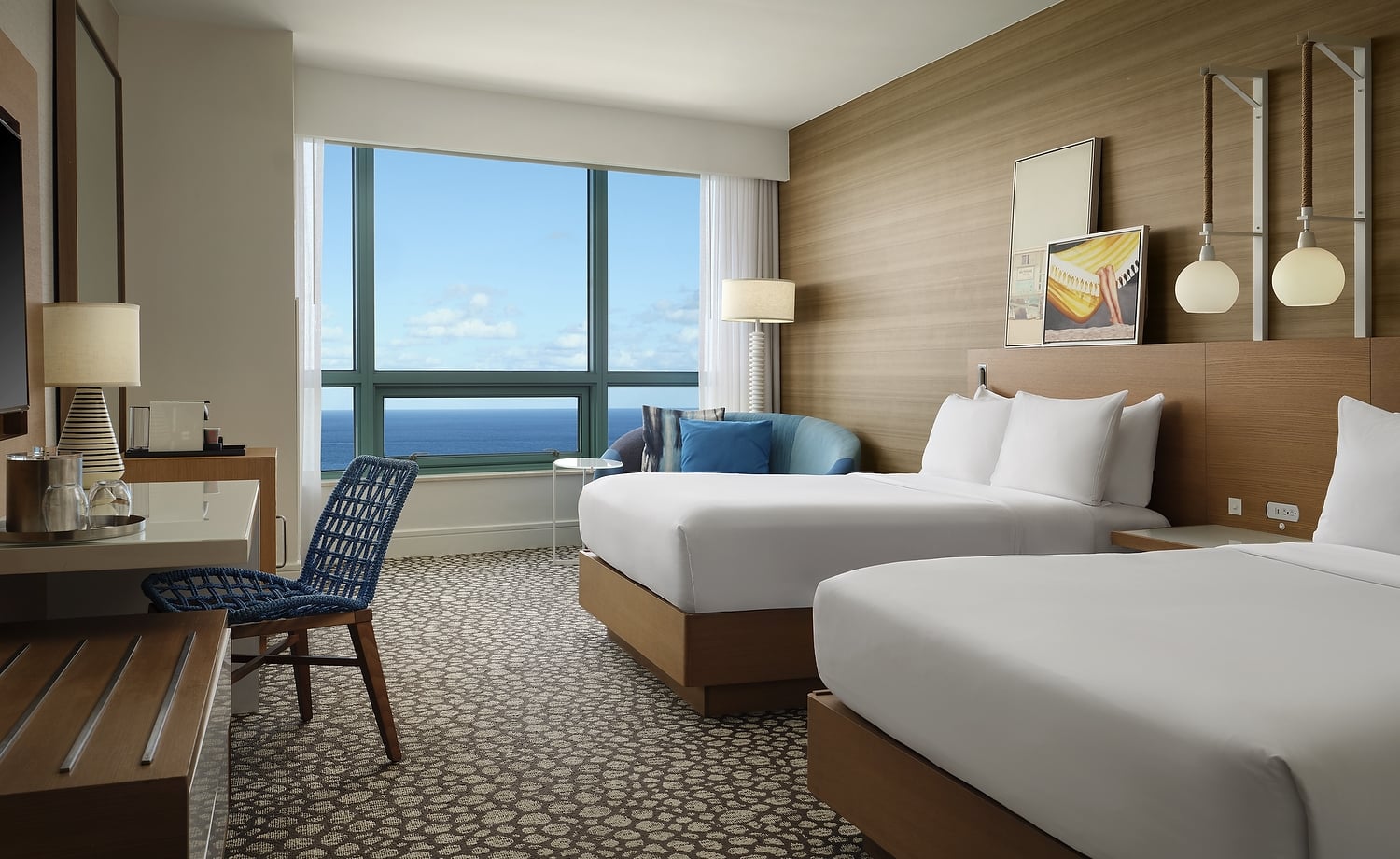 A modern hotel room features two beds, a desk, and a sofa, all arranged to face a large window presenting a vast blue ocean and sky view.