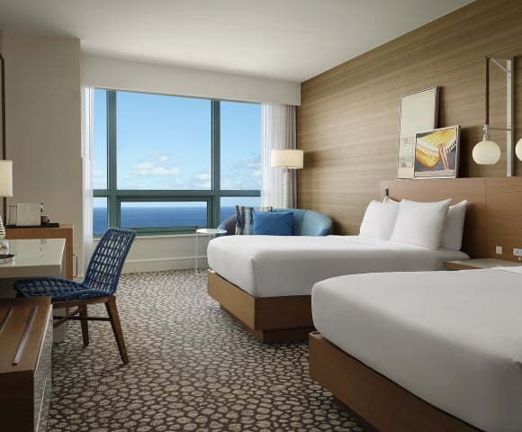 A modern hotel room features two beds, a desk, and a sofa, all arranged to face a large window presenting a vast blue ocean and sky view.