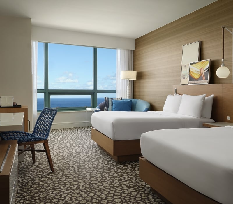A modern hotel room features two beds, a desk, and a sofa, all arranged to face a large window presenting a vast blue ocean and sky view.