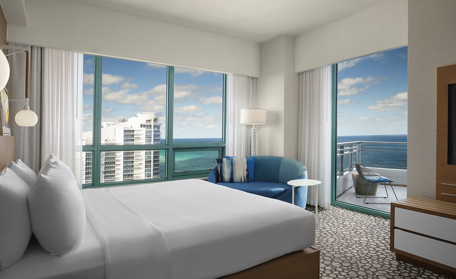 A large bed dominates a modern hotel room, facing expansive windows showing a city skyline and the ocean. A blue sofa and a balcony with an ocean view are also present.