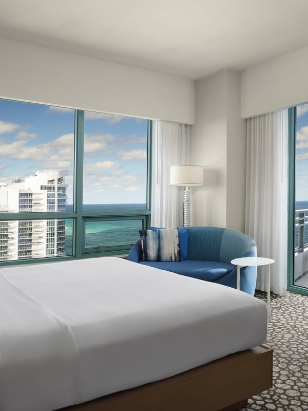A large bed dominates a modern hotel room, facing expansive windows showing a city skyline and the ocean. A blue sofa and a balcony with an ocean view are also present.