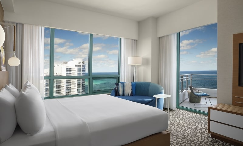 A large bed dominates a modern hotel room, facing expansive windows showing a city skyline and the ocean. A blue sofa and a balcony with an ocean view are also present.