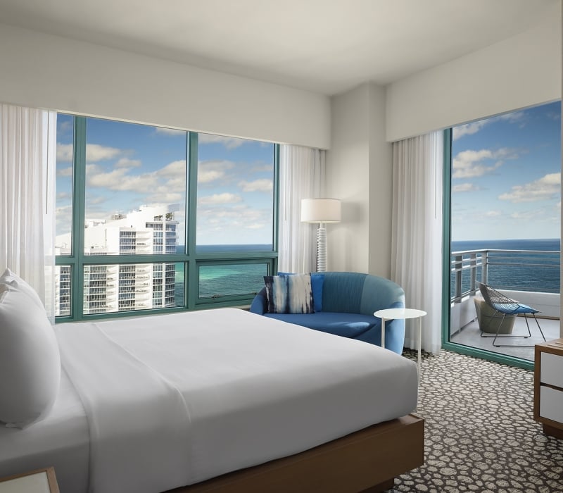 A large bed dominates a modern hotel room, facing expansive windows showing a city skyline and the ocean. A blue sofa and a balcony with an ocean view are also present.
