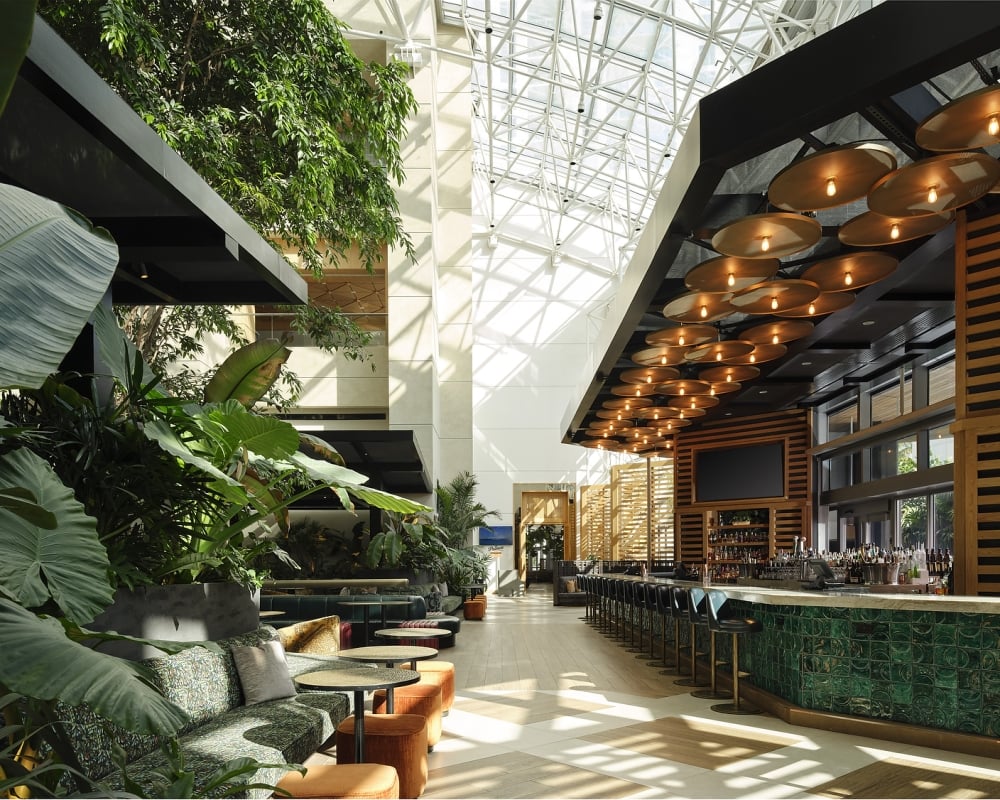 A spacious, modern hotel lounge features a long, green tiled bar and abundant tropical plants providing a lush seating area. Natural light streams from a high glass atrium ceiling. Text: EXIT, Palm Court.